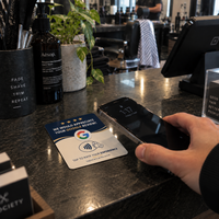 A customer taps their phone on a navy Tap Star card on a dark barbershop counter